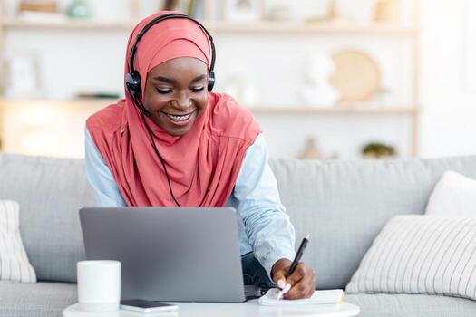 A Black Muslim woman in a hijab wearing a headset, studying on a laptop and taking notes